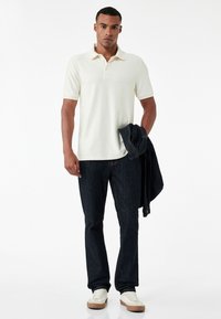 Jasno beżowa koszulka polo z kołnierzykiem i trzema guzikami, zestawiona z ciemnoniebieskimi dżinsami. Model trzyma dżinsową kurtkę. Sneakersy w kolorze kremowym z brązowymi akcentami.