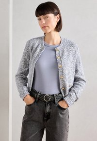 Világoskék tweed kabát kerek kivágással, texturált anyaggal és arany gombokkal. Szürke pólóval és fekete szőtt övvel párosítva.