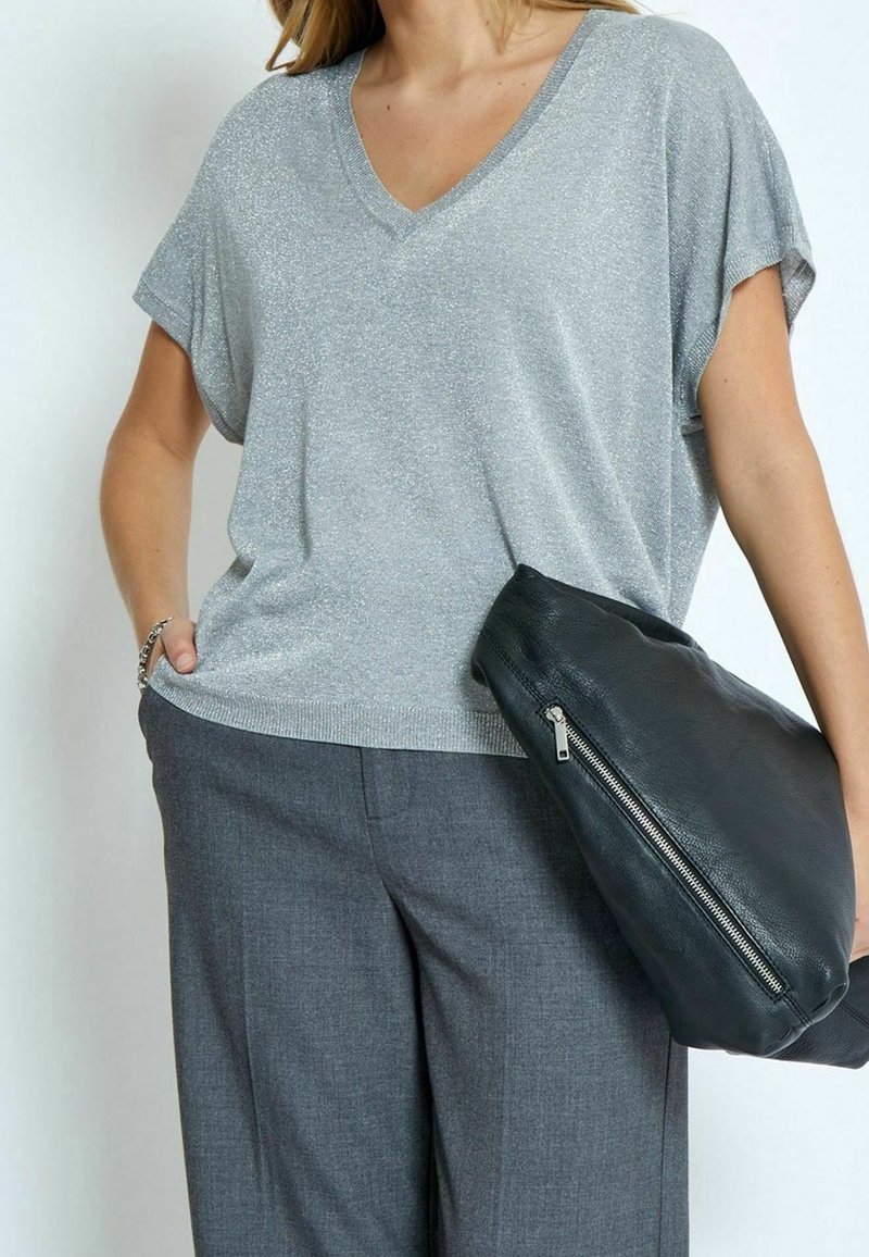 Femme portant un haut argenté à manches courtes et col en V scintillant ainsi qu'un pantalon gris, tenant une grande pochette noire en cuir avec fermeture éclair.