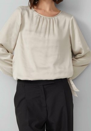 Žena nosící krémovou halenku s dlouhým rukávem a nařaseným výstřihem a černé krejčovské kalhoty, stojící na šedém pozadí.