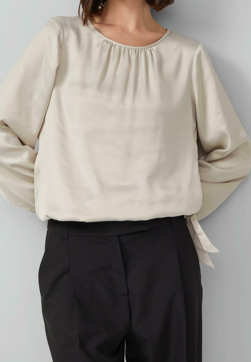 Femme vêtue d'un chemisier crème à manches longues avec un col froncé et d’un pantalon noir sur mesure, debout devant un fond gris.