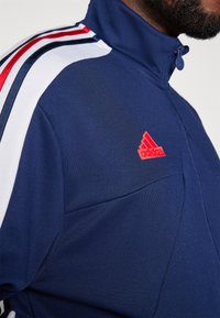 Giacca da allenamento blu navy con zip e dettagli a strisce rosse e bianche. Presenta un logo Adidas ricamato di colore rosso sul lato sinistro del petto.