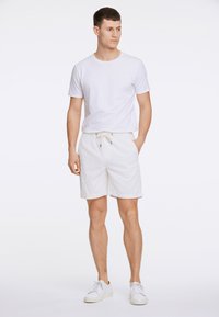 Camiseta blanca de cuello redondo combinada con pantalones cortos de tiro ajustable en beige claro. Los pantalones cortos cuentan con una tela texturizada y bolsillos laterales. Zapatillas blancas.