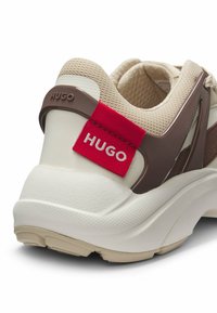 Hugo sneakers feature a beige mesh upper, brown accents, and a white textured sole. A red tag with "HUGO" is prominently displayed at the heel.