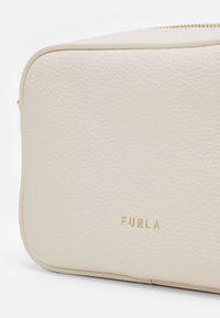 Bílá kosmetická taška z pebble kůže s zlatým logem "FURLA", obdélníkový tvar, hladká textura a zipové uzavření nahoře.