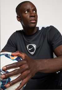 Czarna koszulka sportowa z białymi akcentami, z logo Nike na piersi, teksturowany materiał. Model trzyma białą piłkę nożną w niebieskie wzory.