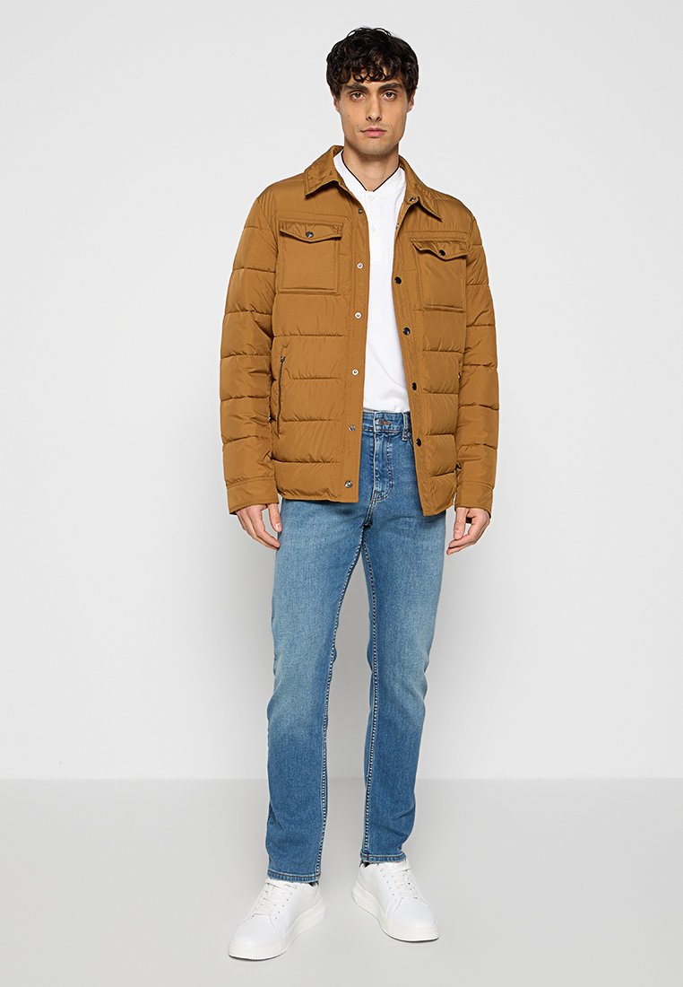 Homme portant une veste matelassée marron, une chemise blanche, un jean bleu et des baskets blanches, se tenant devant un fond blanc uni.