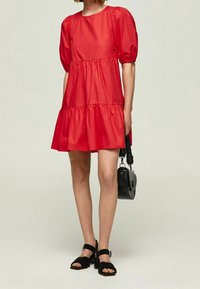 Vestido de algodón rojo con mangas abullonadas, diseño en capas y un escote redondo. Combinado con sandalias negras y un pequeño bolso negro.