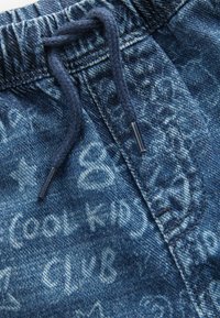 Denimist joggers paelaga, millel on heledam sinine värv ja käsitsi kirjutatud valged tekstitooted, mis katab kogu kanga.
