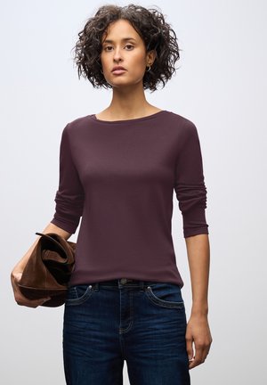 Bordeaux lange mouwen top met een ronde hals, gladde textuur, gecombineerd met donkerblauwe jeans. Het model houdt een bruine tas vast.