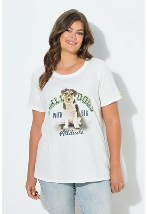 Smilende kvinde iført hvid T-shirt med et lille hundebillede og teksten "Små hunde med stor attitude", kombineret med lyseblå jeans.