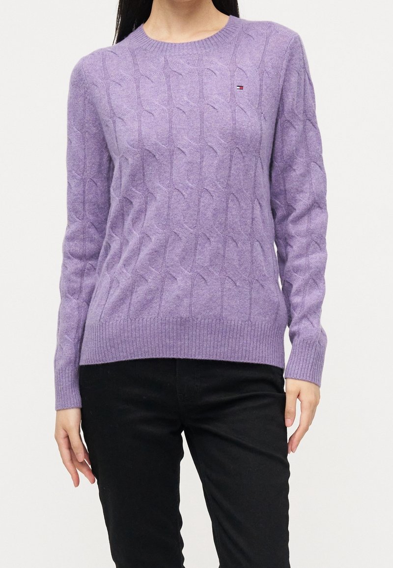 Femme portant un pull en maille torsadée lavande avec logo sur la poitrine et un pantalon noir, debout devant un fond clair uni.