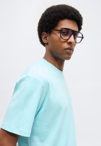 Νέος άνδρας με κοντά σγουρά μαλλιά που φοράει μεγάλα σκουρόχρωμα γυαλιά με σκελετό και ανοιχτό μπλε μπλουζάκι, κοιτάει ελαφρώς πλάγια.
