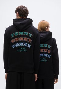 Μαύρα φούτερ με κουκούλα με μεγάλο πολύχρωμο κείμενο "TOMMY" και εκτύπωση "JEANS New York City" στο πίσω μέρος. Απλός σχεδιασμός, βαμβακερό υλικό.