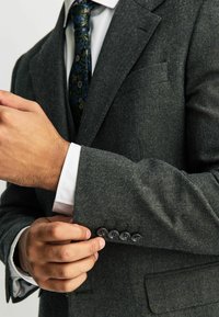 Blazer de lana gris carbón con una textura sutil y cinco botones oscuros en las mangas. Camisa blanca con un patrón de rayas y corbata estampada visible.