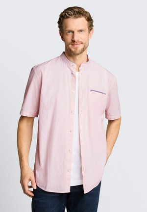 Man met lichtbruin haar en baard, die een lichtroze kort mouwen overhemd draagt over een wit T-shirt en een donkere broek, staand tegen een effen achtergrond.