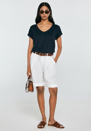 Kobieta ubrana w czarny T-shirt z dekoltem w serek, białe szorty z paskiem, brązowe sandały, okulary przeciwsłoneczne, złote kolczyki koła i trzymająca małą torebkę.