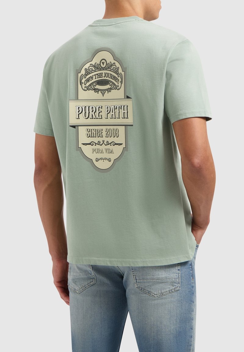 Lichtgroene katoenen t-shirt met een grote grafische afbeelding in crème, tekst "PURE PATH" met een sierlijke bannerontwerp op de achterkant. Casual fit.