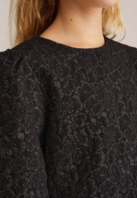 Vestito nero con motivo floreale testurizzato, scollatura tonda e leggero volume sulle spalle. Il tessuto sembra ricco e stratificato.