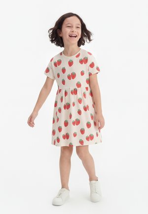 Niña sonriente con cabello corto y oscuro, vestida con un vestido blanco con estampado de fresas rojas y zapatillas blancas, caminando sobre un fondo blanco.