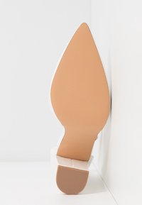 Semelle d'une chaussure à talon haut à bout pointu avec un dessous lisse beige et un talon carré texturé, positionnée verticalement contre une surface blanche.