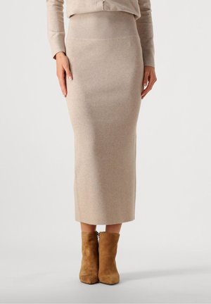 Kvinde iført en beige ribstrikket midi-nederdel kombineret med matchende top og lysebrune ruskindsstøvler, stående mod en ensfarvet baggrund.