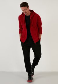 Crvena zip-up dukserica s mekim teksturom i dva prednja džepa, uparena s crnim hlačama i crnim visokim tenisicama s crvenim akcentima.