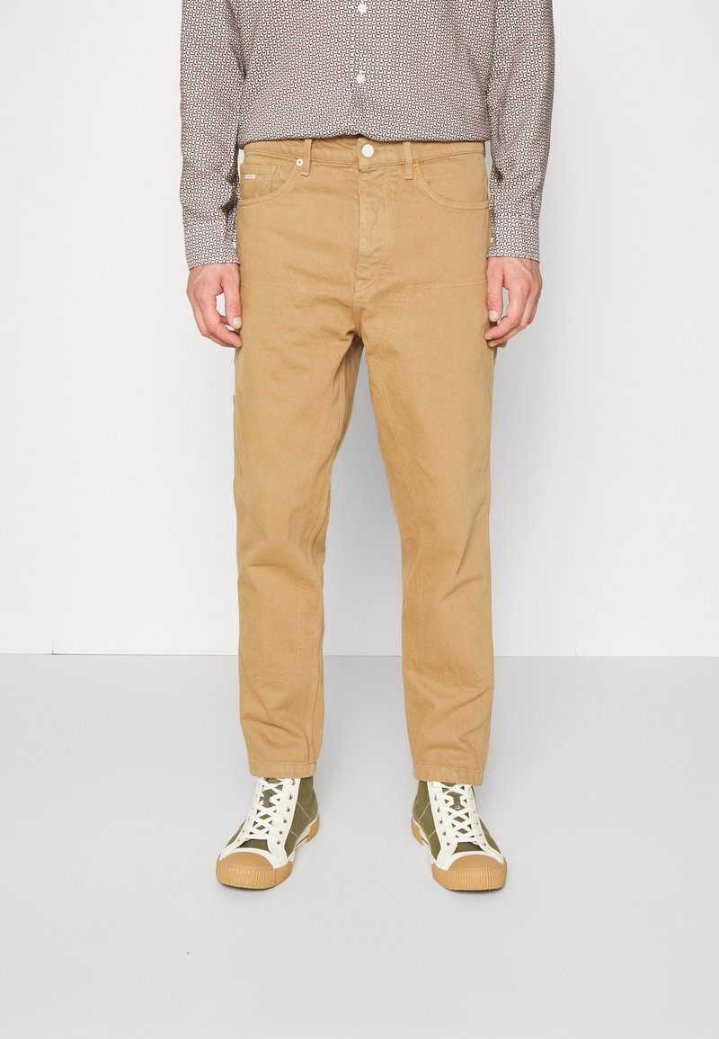 Pantalon en velours côtelé beige à coupe droite, doté de poches avant et de coutures visibles. Porté avec des baskets montantes vertes.