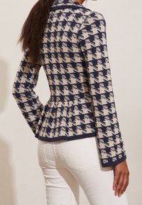 Stickad jacka med ett midnattsblått och krämfärgat hundtandsmönster, peplummidja, långa ärmar och en mjuk textur.