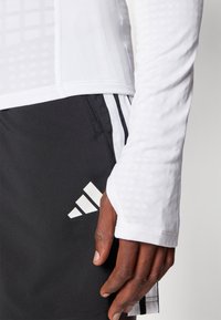 Weißes langärmliges Sportoberteil mit strukturiertem Design, kombiniert mit schwarzen Shorts, die ein weißes Adidas-Logo und Seitenstreifen zeigen.