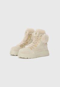 Bottines beige à lacets avec un col en fourrure et des accessoires en or, placées sur un fond blanc uni.
