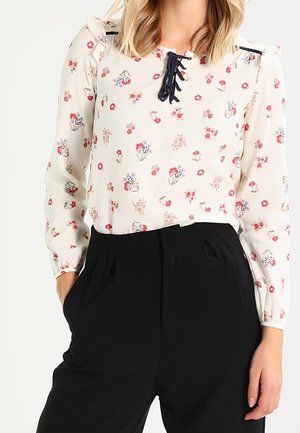 Femme portant un chemisier crème à petits imprimés floraux roses et rouges, avec un détail en dentelle noire à lacets, associé à un pantalon noir taille haute.