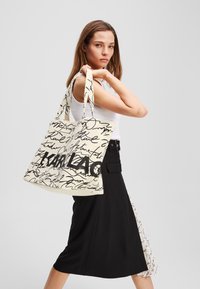 Bolsa de lona con fondo crema y texto en estilo manuscrito negro que incluye el logo "KARL LAGERFELD". Con asas largas para facilitar el transporte.