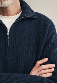 Pulover din tricotaj bleumarin cu guler cu fermoar. Textură groasă, detalii de cusătură vizibile și o buzunară subtile în față, aproape de guler.