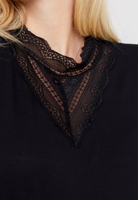 Svart topp med en spetskrage med intrikata mönster, som tillför textur och detalj till halslinjen. Tyget verkar mjukt och slätt.