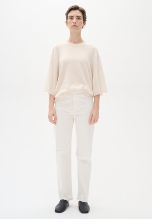 Hellbeige, übergroßes Strickoberteil mit kurzen Ärmeln, kombiniert mit figurschmeichelnden weißen Hosen und schwarzen Slippers. Minimalistisches Design, glatte Textur.