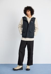 Colete puff preto com bolsos frontais duplos por cima de uma hoodie bege, combinado com calças largas pretas e sapatos de exterior bege.