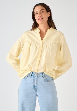 Femme debout, les mains dans les poches arrière, portant un chemisier jaune pâle brodé et un jean taille haute bleu clair.