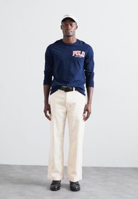 Námořnické tričko s dlouhým rukávem a logem "POLO" v červené barvě, v kombinaci s krémovými pruhovanými širokými kalhotami a černými botami, na hlavě má bílou čepici.