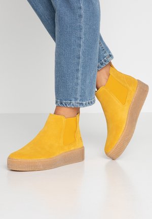 Fødder iført skarpe gule ruskind ankelstøvler med tykke beige såler, kombineret med blå denim cropped jeans mod en hvid baggrund.