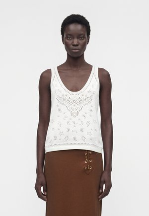 Mujer con cabello corto y rizado que lleva una camiseta blanca sin mangas con patrones recortados y una falda marrón con detalle de cordones en el lateral.