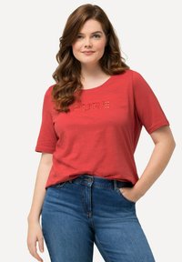 Camiseta roja de manga corta con cuello redondo, que presenta letras recortadas que dicen "PURE" en la parte frontal. Combinada con jeans de mezclilla azules.