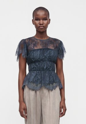 Femme aux cheveux courts portant un haut en dentelle bleu foncé avec des manches transparentes et un pantalon beige à jambes larges sur un fond blanc uni.