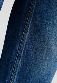 Jeans di denim blu scuro con una texture liscia, caratterizzati da cuciture visibili di un colore arancione contrastante lungo la cucitura laterale.