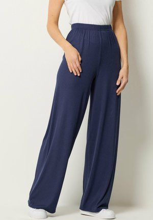 Kobieta ubrana w granatowe, szerokie spodnie z prążkowanego materiału, białą bezrękawnikową bluzkę i białe trampki, stojąca na tle jasnego tła.