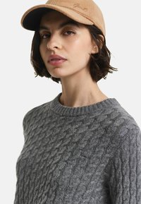 Grau gestrickter Pullover mit Kabelmuster und rundem Ausschnitt. Die Person trägt eine beige Mütze mit gesticktem Text auf der Vorderseite.