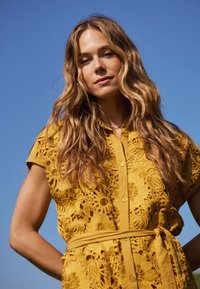 Abito giallo in pizzo con maniche corte, motivo floreale, chiusura con bottoni e cintura staccabile; contro un cielo blu limpido.