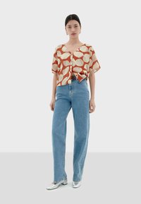 Kurzärmelige, gemusterte Bluse in Rost und Creme, kombiniert mit hellblauen Jeans. Das Model trägt silberne, viereckige Absatzschuhe.