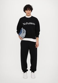 Der schwarze Sweatshirt hat den Aufdruck "No Problemo" in weißer Schrift und wird mit schwarzen Sweatpants und weißen Sneakern kombiniert. Das Model hält gefaltete Jeans.