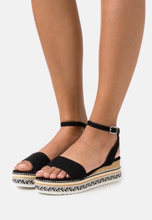 Pieds chaussés de sandales à plateforme à bride cheville noires avec des détails tissés multicolores sur la semelle épaisse sur un fond uni.